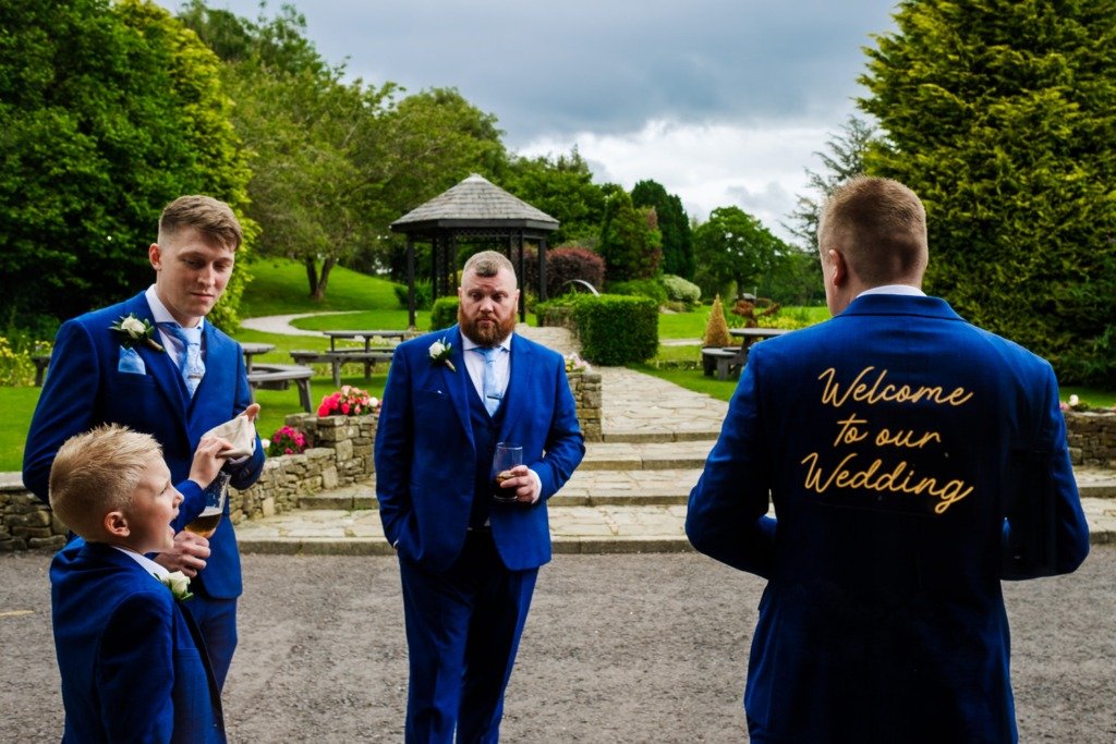 The groom, with a reflection of a wedding sign on his back, and his guests stand outside at Mytton Fold Hotel