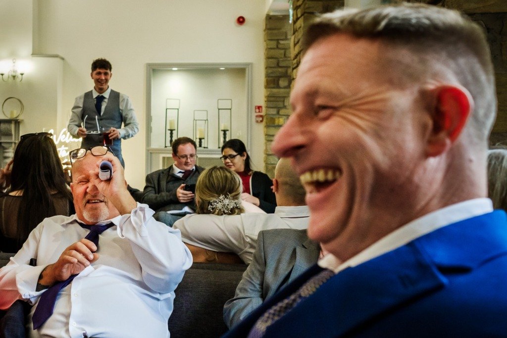 A guest laughs as another guest looks through a rolled-up piece of paper at a Mytton Fold wedding in the Ribble Valley