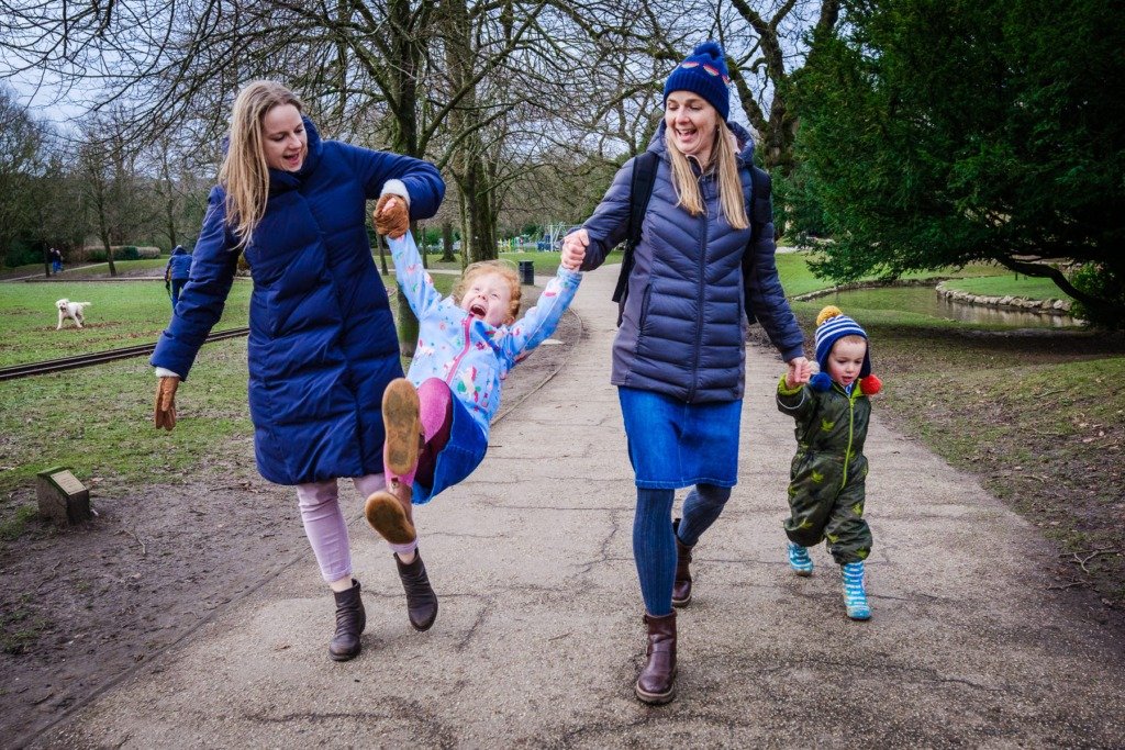 Two adults swing a laughing child during a Buxton family photography session in Pavilion Gardens